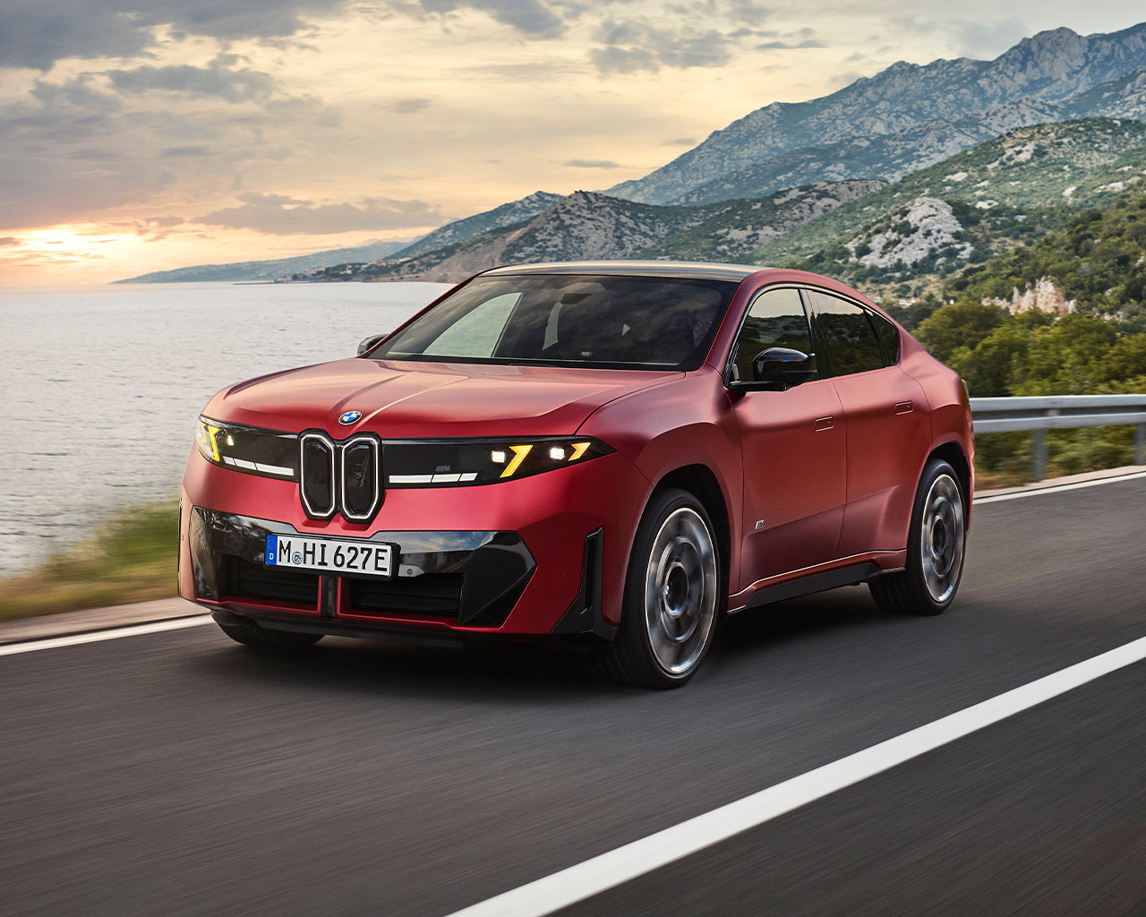 A red BMW iX4 M60 xDrive seen from the front left drives on a coastal road at dusk, with the sea and mountains in the background.