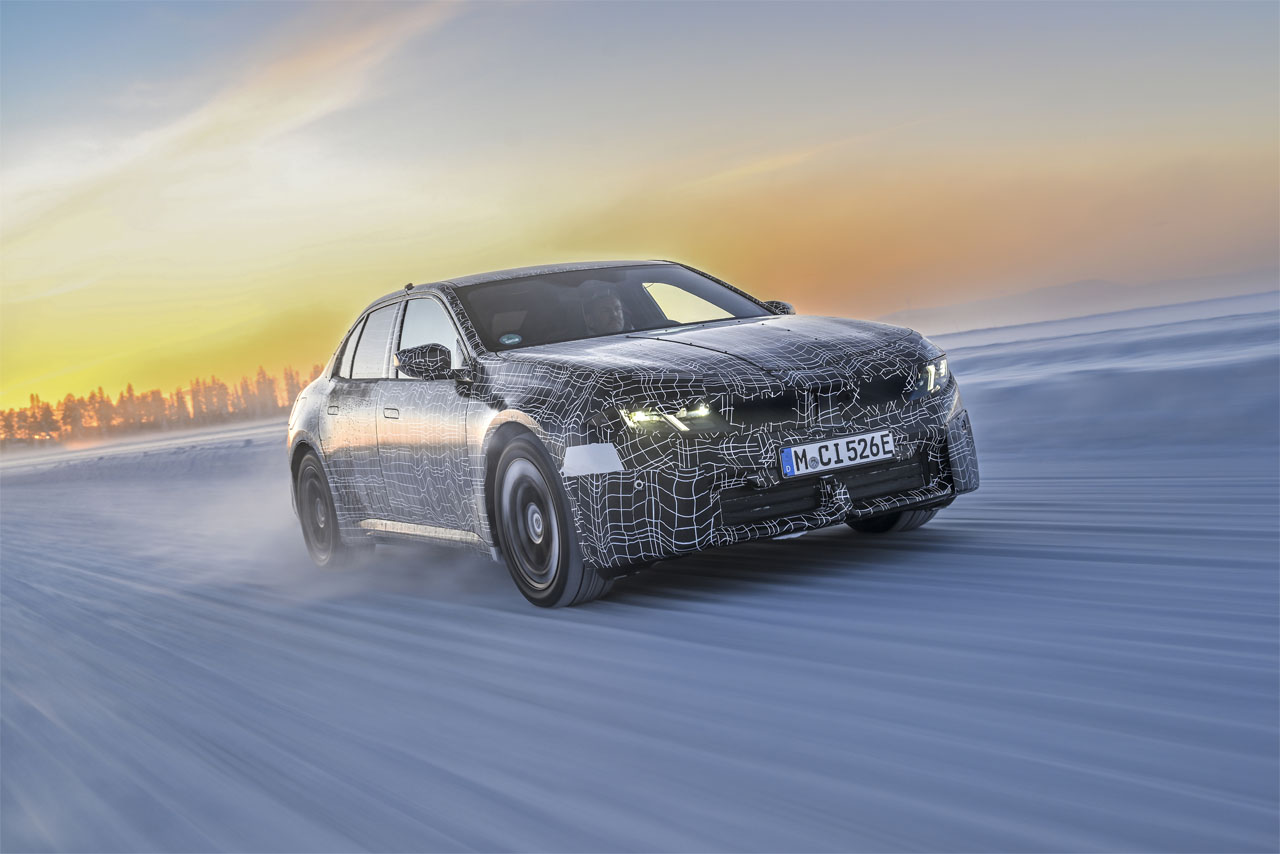 BMW electric car with camouflage foil drives on a snowy road at sunset.
